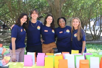 Three full-time staff and two undergraduate student workers in navy NDatUT shirts stand smiling in front of a table with a prize-wheel and multi-color giveaway bags with fidgets.