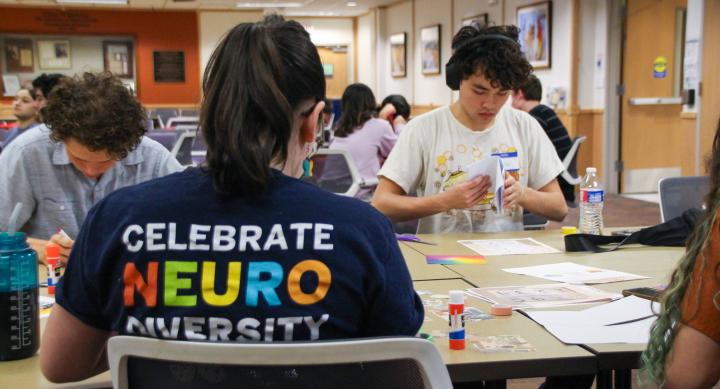 Two students crafting at a table next to a student worker wearing a shirt that says "Celebrate Neuro Diversity"